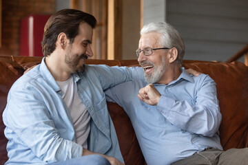 Excited mature father in glasses with grown-up son talking, having fun, hugging and sitting relaxing on cozy couch at home together, smiling young man with senior dad two generations enjoying weekend