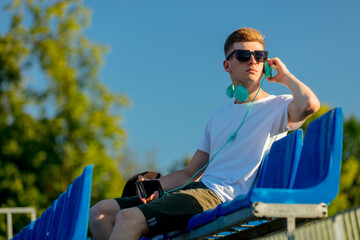 Young teen boy in sunglasses with headphones sitting in blue sit on stadium tribune