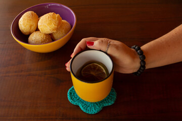 Uma mão feminina segurando uma caneca de chá e uma vasilha de pães de queijo.