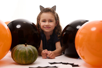Fototapeta premium Smiling adorable child girl in carnival costume, little witch lying down on white background with copy space next to pumpkins, handmade bats and colorful orange black balloons. Halloween party concept