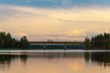 Fototapeta premium Landscape with a river and a forest illuminated by the setting sun. Reflection of clouds in the water. Suburban road bridge through the river.