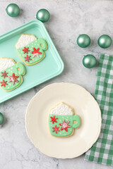 Homemade Christmas Cookies Shaped Like Coffee Mugs on White Background with Christmas Ornaments, Green Checked Towel, Green Cookie Sheet, One on White Plate