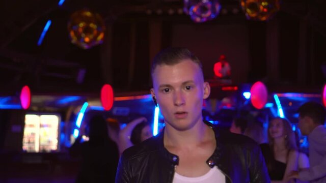 A guy walks through a nightclub right in front of the camera. Movement. He is wearing a black jacket and dilated black pupils in his eyes. On the background of the dance floor and dancing people.