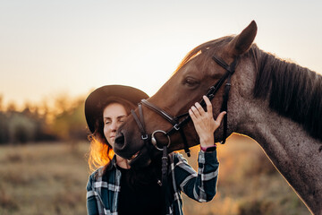 Beautiful smiling young woman in hat and shirt playing with brown horse in a field at sunset. Horseback riding.