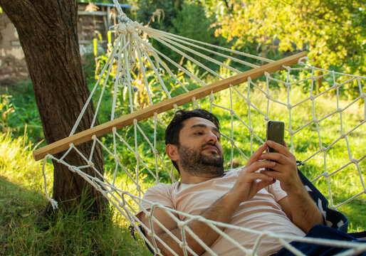 Man looking at his phone while lying in a hammock in the garden of his house.