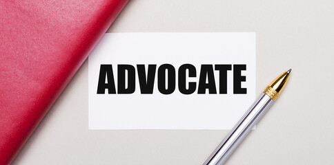 On a light gray background, there is a golden pen, a burgundy notebook and a white blank card with a place to insert text ADVOCATE. Business concept. Template