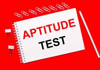 On a bright red background, a white pencil, white paper clips, and a white notebook with the text APTITUDE TEST