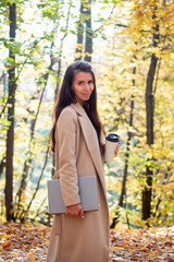 Brunette woman in autumn park with laptop and cup of coffee. Student. Teacher. Online education.