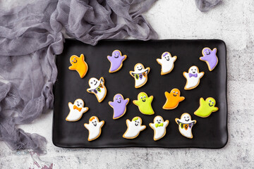 Obraz premium Homemade Halloween Cookies Sahped Like Ghosts and Decorated with White, Purple, Green, and Orange Icing; Gray Cheesecloth Placed Beside Black Plate on Light Gray Background; Top View