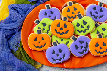 Homemade Halloween Cookies Decorated Like Orange, Purple, and Green Pumpkins; Arranged on a Pumpkin Shaped Plate; Yellow, Purple and Green Cheesecloth Beside; Light Gray Cement Background
