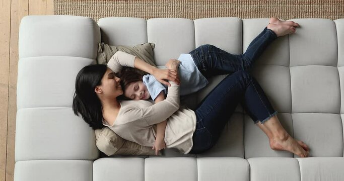 Above top view affectionate caring young korean asian mother cuddling little cute adopted kid son, sleeping together on huge cozy couch, watching sweet dreams, relaxing on carefree weekend at home.