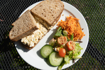 Fresh sourdough bread egg sandwich with a side salad of cucumbers, tomatoes, lettuce, carrots and spring onions served on white plate.