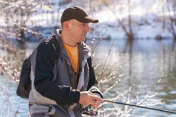 A fisherman with a fishing rod and a backpack catches fish on the bank of a snow-covered river in early spring