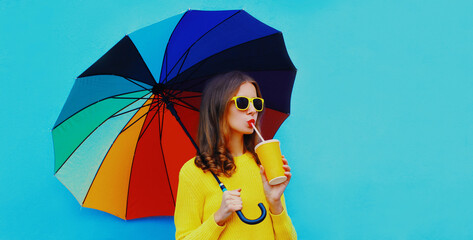 Autumn portrait of young woman with colorful umbrella drinking a juice wearing an yellow knitted sweater on blue background © rohappy