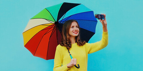 Portrait of happy smiling young woman taking a selfie picture by smartphone with colorful umbrella on blue background
