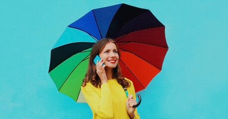 Portrait of smiling young woman calling on smartphone with colorful umbrella on blue background