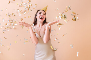 Fototapeta premium Happy woman in party hat and with flying confetti on color background