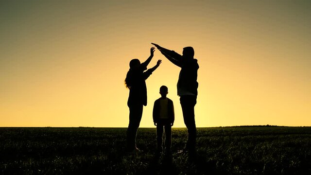 Children mom and dad are playing together building house with their hands, silhouette at sunset. Happy family dreams of their own home in park in summer in sunshine. Taking care of children by parents