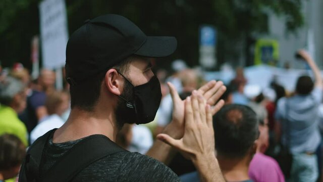 Young adult protester man riot guy in covid-19 facemask claps on protest picket. Male activist in black coronavirus face mask applauds. Rebels on revolted strike, standing demonstration rally crowd.