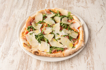 Small Italian pizza with mozzarella cheese, Parmesan cheese slices, arugula leaves, prosciutto slices and olive oil on wooden table