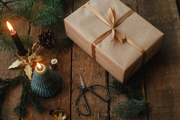 Stylish christmas gift wrapped in craft paper, candle, scissors, fir branches on rustic wood....