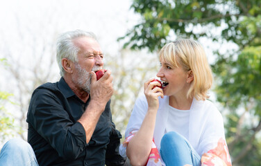 Senior caucasian couple sitting on field grass and enjoy eating apple together outdoors in parks. Happy mature couple eating fruits in a summer park. Healthy and lifestyle retired couple outdoors.