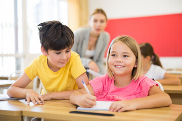 Portrait of small school girl and boy sitting together in classroom during lesson in elementary school