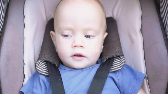 cute and nice blue eyed caucasian baby infant fastened with black seatbelts in brown kid carriage. lying in stroller moving his leg and looking straight into camera and around with big eyes