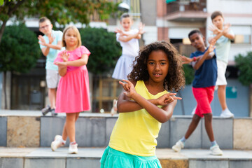 Smiling cute african american preteen girl dancing modern choreography with group of tweenagers on city street on summer day
