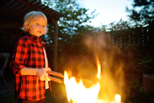 Cute Little Boy Is Burning A Bonfire On A Summer Evening In The Backyard. Child Puts Firewood In A Fire Bowl By Tongs. Summer Holidays In Countryside.