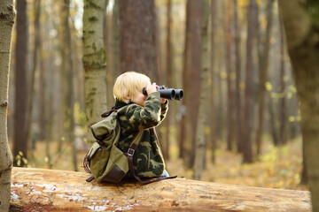 Little boy scout with binoculars during hiking in autumn forest. Child is sitting on large fallen tree and looking through a binoculars. Adventure, scouting and hiking tourism for kids.