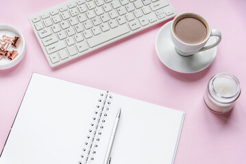 Cozy workplace with blank notepad, keyboard, cup of coffee and candle. Top view, flat lay. Pink background