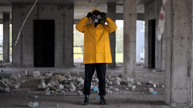stalker, survivor, stands in yellow raincoat and black gas mask in abandoned building and wears hood over his head. Post-apocalypse concept, biochemical threat, post-covid-19.