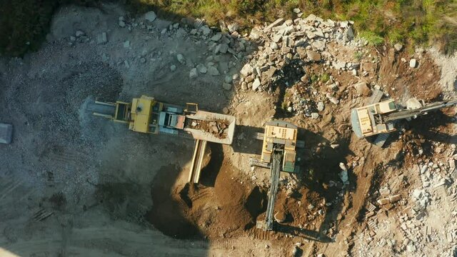Drone shot of two Excavators loading into a transportable industrial stone crusher with granite for gravel production at a construction site. Heavy excavators throw large rocks with buckets and smash 