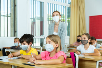 Fototapeta premium School boy and girl in face masks listening to teacher and writing exercises in notebook at lesson in primary school