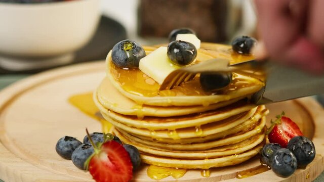 Cutting pancakes with berries using knife and fork close-up. Eating fresh cooked blueberries and strawberries flapjacks for breakfast. Stack of oatmeal pancakes. Healthy vegetarian sweet food. 