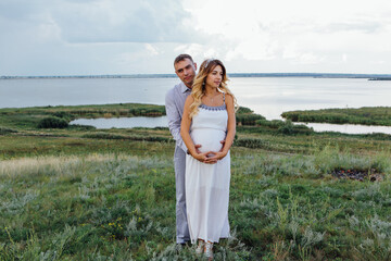 Happy young couple, expecting a baby. Man hugs his pretty pregnant woman in a white dress outdoors.