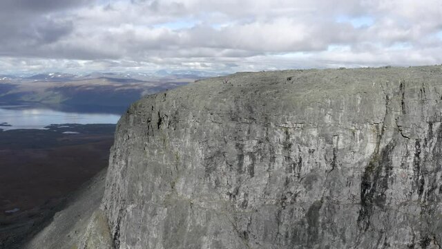 An aerial drone shot circling a large cliff with a small group of people on top looking over a vast cloudy landscape - daytime