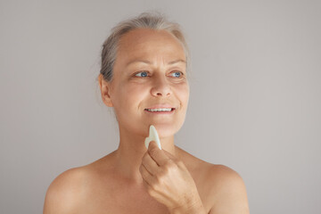 Senior naked woman massages her face with a jade board. Isolated on gray background.