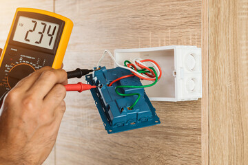 Close up hand of an electrician uses a digital meter to measure the voltage at a wall socket on a wooden wall.