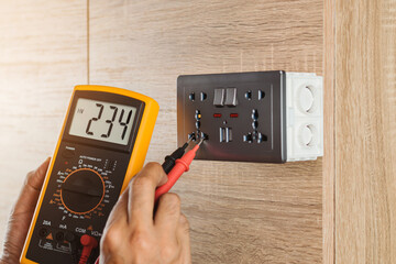 Close up hand of an electrician uses a digital meter to measure the voltage at a wall socket on a wooden wall.