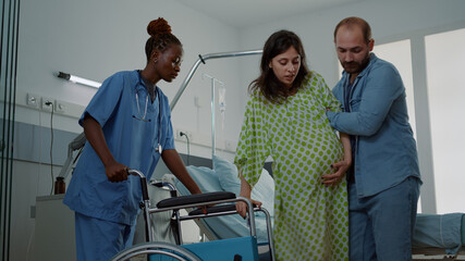 Caucasian man helping pregnant woman in hospital ward and african american nurse bringing...