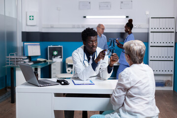 Medic of african american ethnicity holding x ray scan explaining disease to elder woman in cabinet. Black man and old patient doing checkup while nurse and senior man talking in background