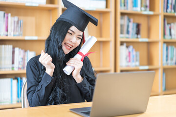 Young Asian university woman graduate in graduation gown and mortarboard with degree certificate celebrates with family through video call on laptop in virtual convocation during COVID-19 pandemic