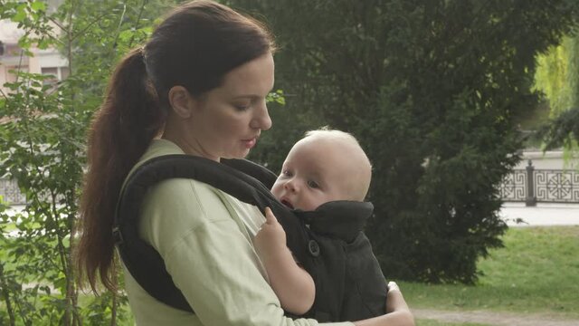 Mother carrying adorable little baby with ergonomic infant kid carrier and walking in public park, six month old baby boy in baby hipseat sling on a walk outdoors. High quality 4k footage