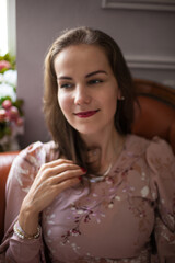 Naklejka premium Young beautiful woman in pink dress and stylish accessories sits on brown leather sofa on white wall background. Portrait of happy smiling girl with red lips resting at home and looking at camera. 