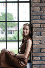 Beautiful young woman in brown leather dress resting on the windowsill. Portrait of happy smiling girl sits on the stained glass window at industrial loft wooden brick style interior. Leisure concept.