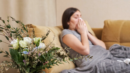 Allergic lady with long hair wipes nasal spray with paper napkin and applies medicine sitting on brown sofa near bouquet of flowers at home