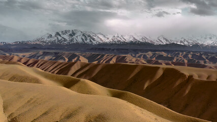 A unique landscape of wild nature with a valley of yellow sand dunes against the background of a large rocky mountain with peaks covered with white snow. A wonderful landscape of environment.