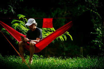 asian old man sitting in red hammock and looking to his mobile phone in the morning